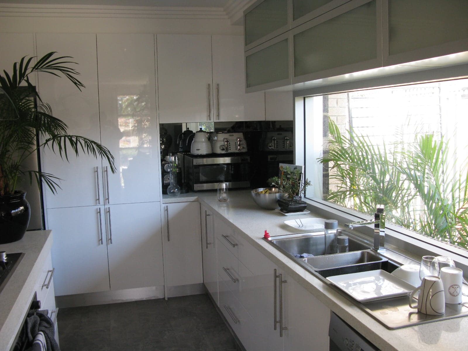 A bright, high-gloss white kitchen with mirrored splashback, stainless appliances and a garden-view window, completed by Freedom Cabinet Australia